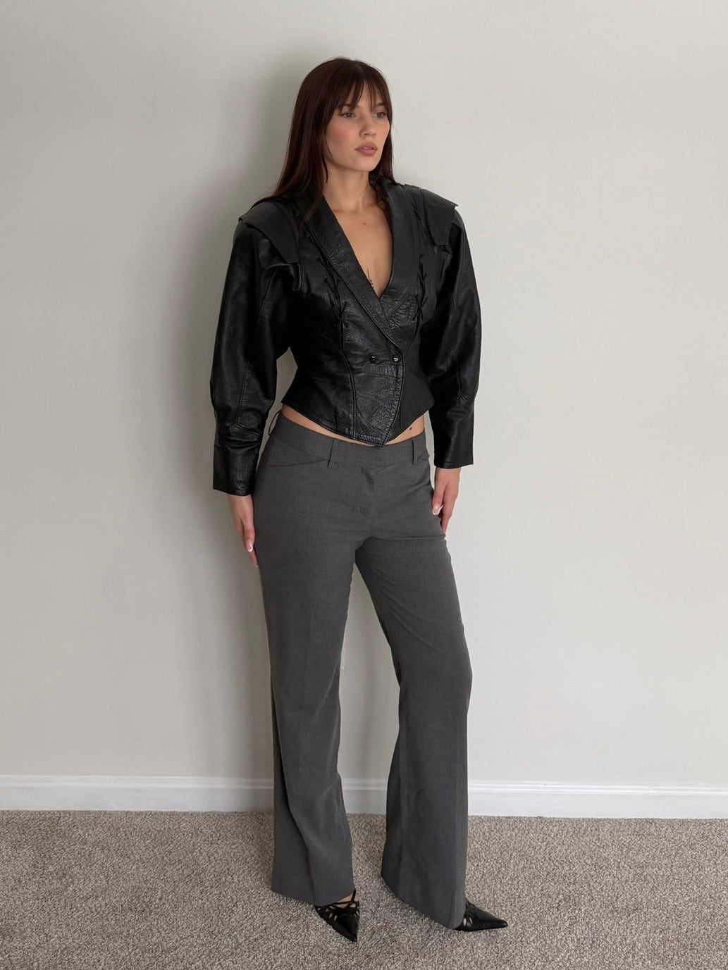 Woman wearing a black leather jacket and gray pants against a plain wall.