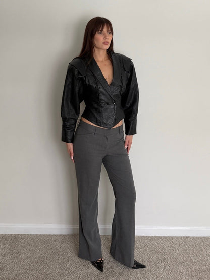 Woman wearing a black leather jacket and gray pants against a plain wall.