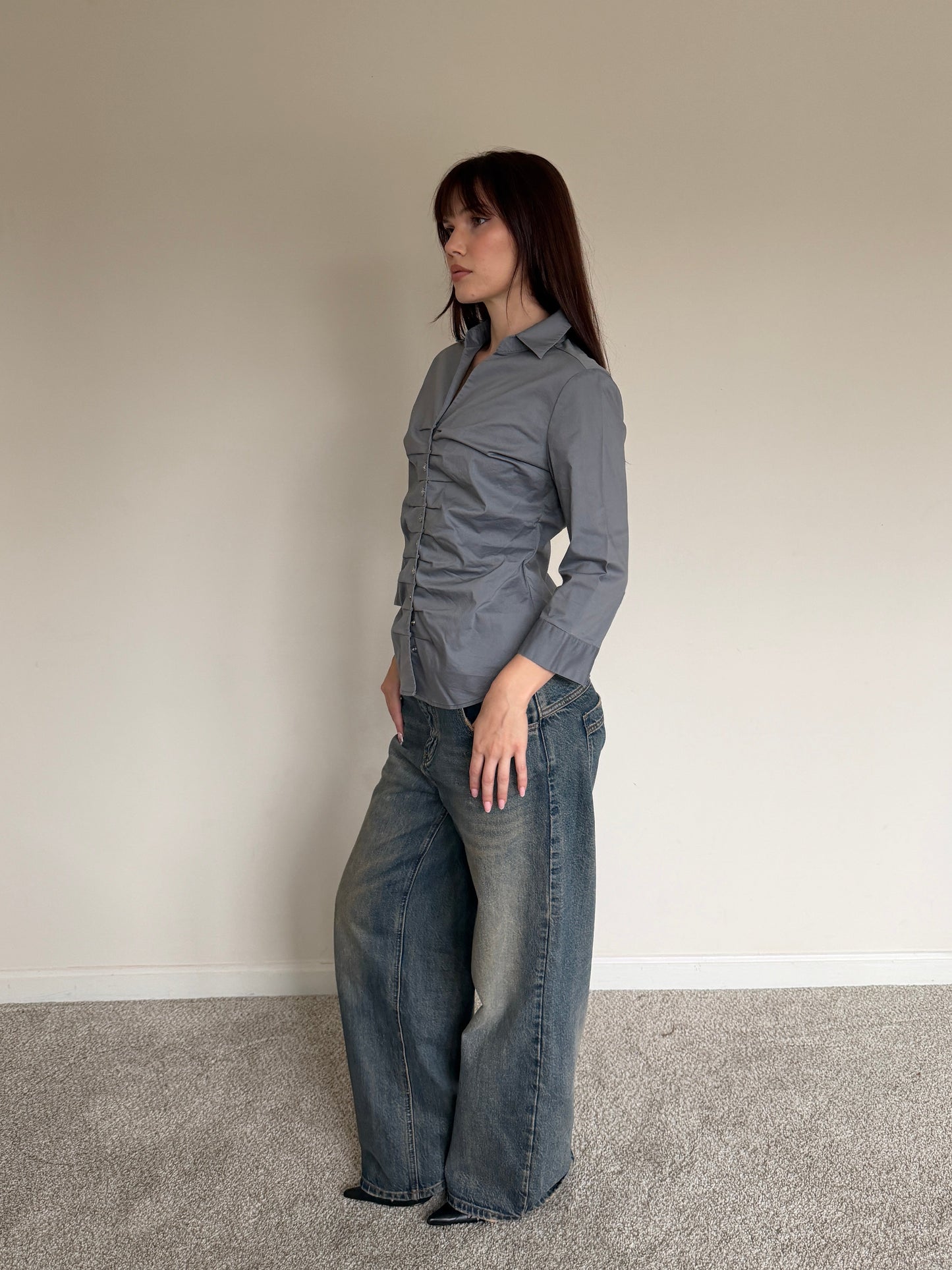 Woman wearing a gray shirt and blue jeans standing against a plain wall.