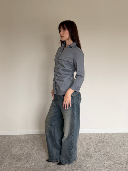 Woman wearing a gray shirt and blue jeans standing against a plain wall.