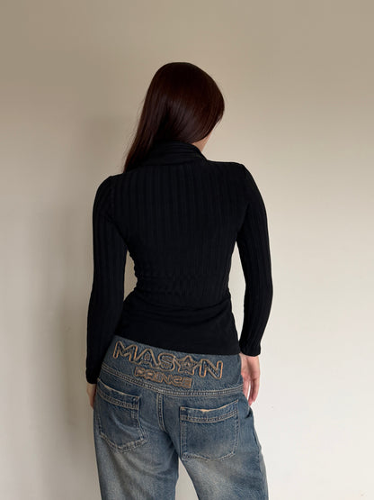 Person wearing a black long sleeve and blue jeans, standing against a plain wall.