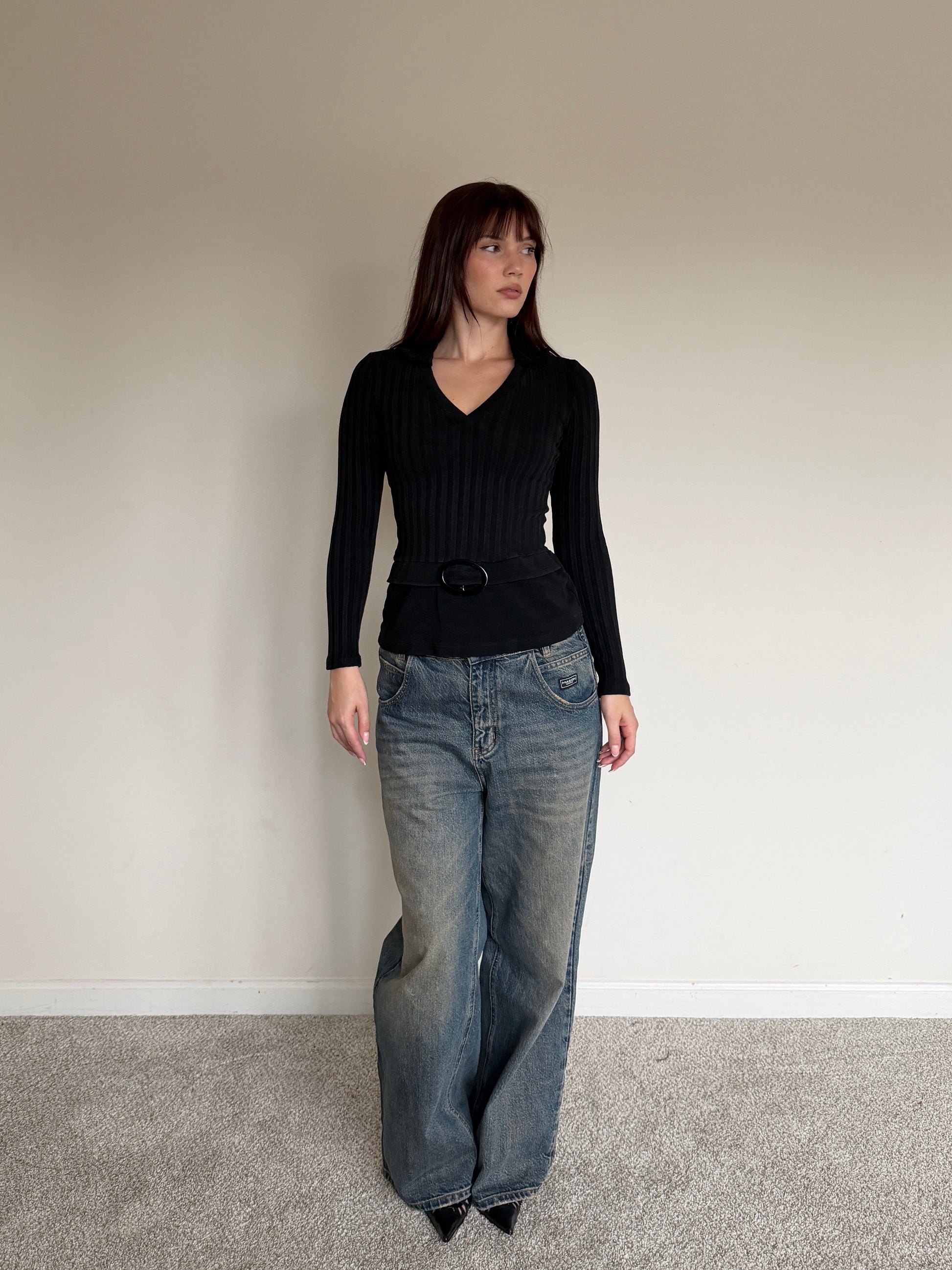 Woman wearing a black top and blue jeans standing against a plain wall.