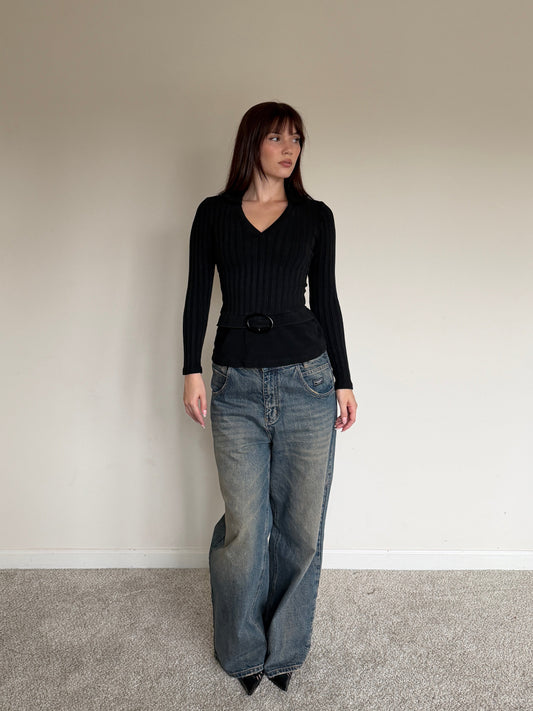 Woman wearing a black top and blue jeans standing against a plain wall.