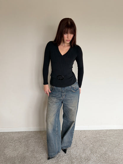 Person wearing a black top and blue jeans against a plain wall.