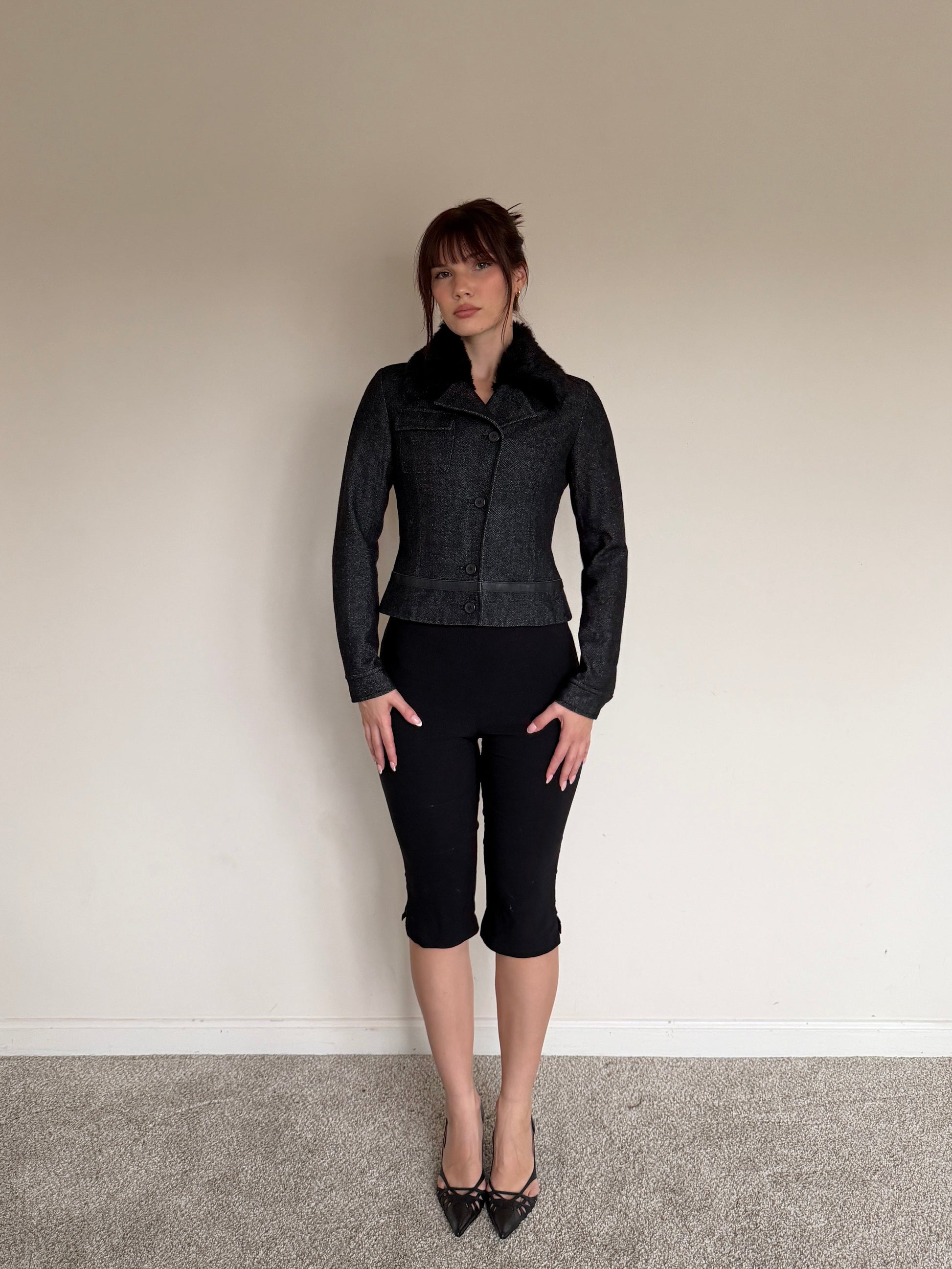 Person wearing a black jacket and black pants standing against a plain wall.