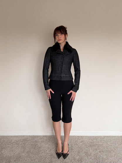 Person wearing a black jacket and black pants standing against a plain wall.