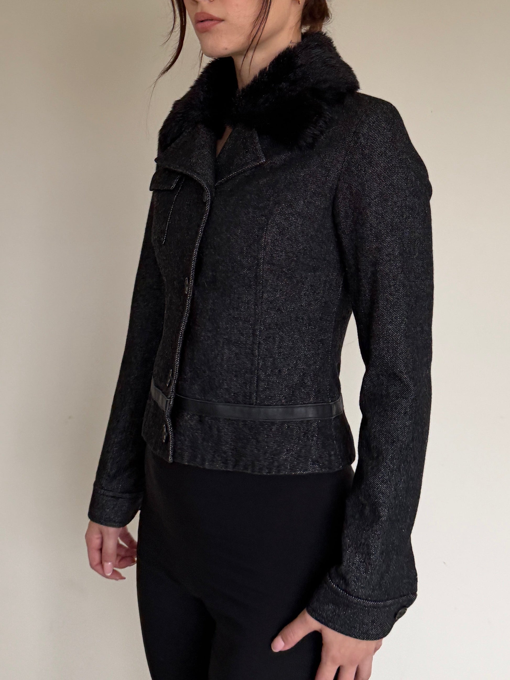 Woman wearing a dark gray jacket with a fur collar against a plain wall.