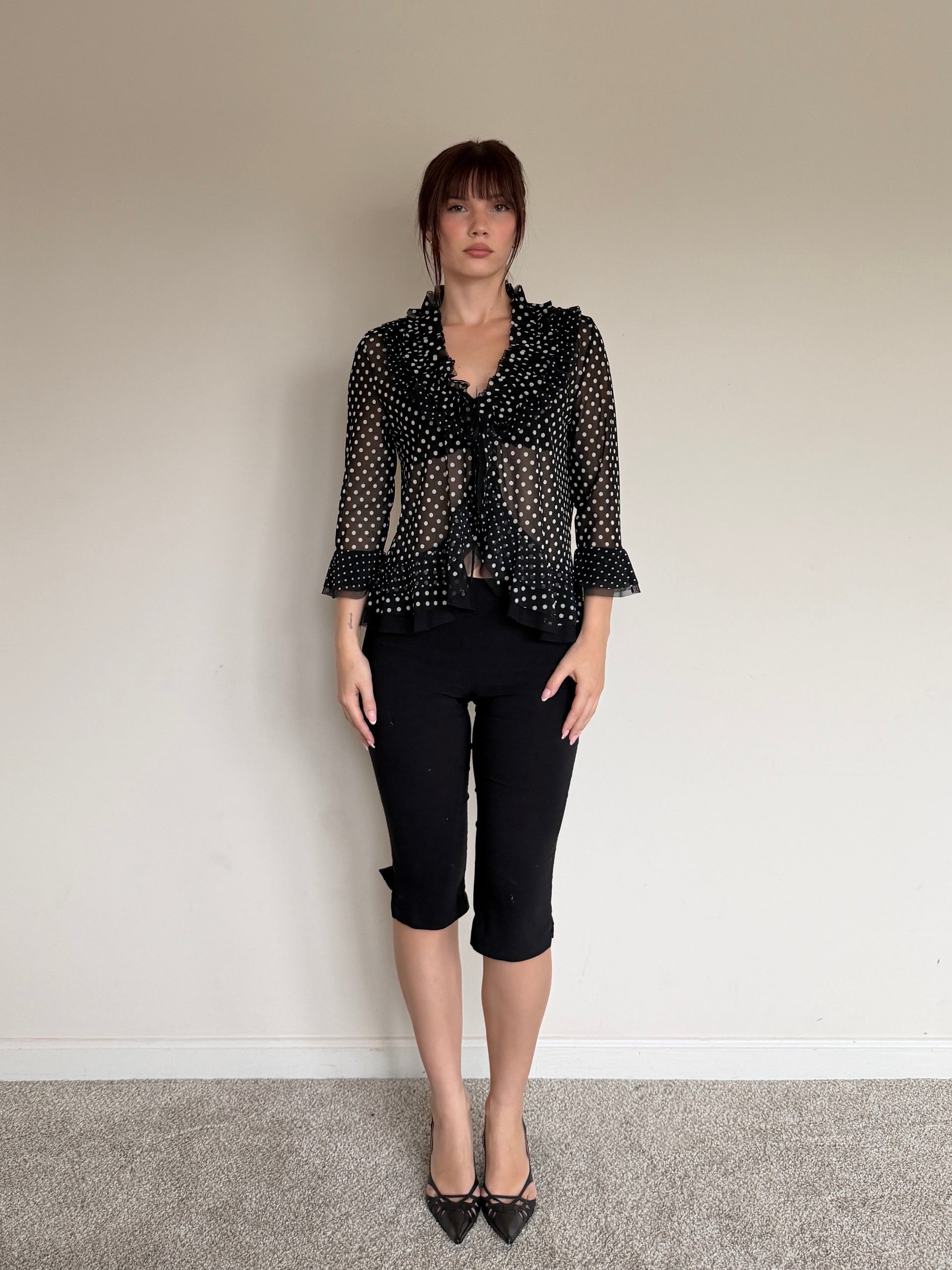 Person wearing a black polka dot blouse and black pants against a plain wall.