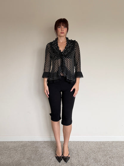 Person wearing a black polka dot blouse and black pants against a plain wall.