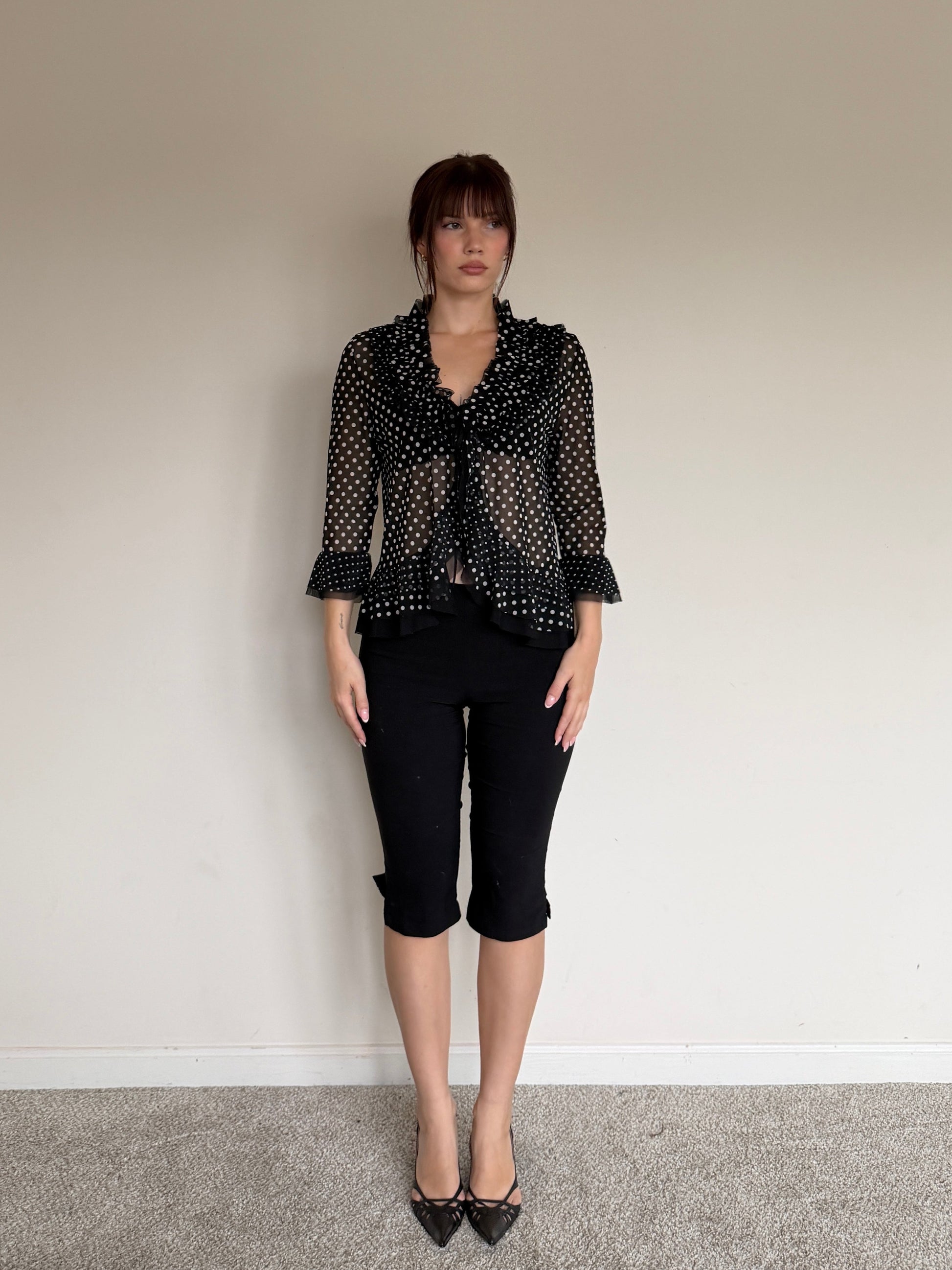 Person wearing a black patterned blouse and black pants against a plain wall.