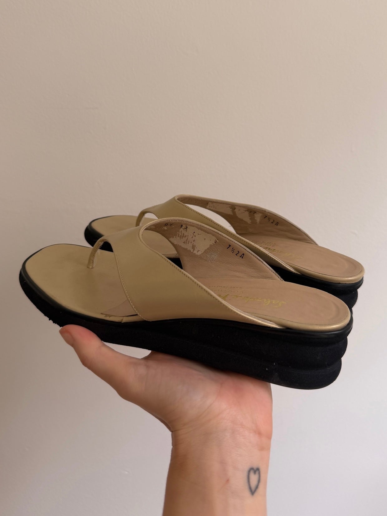 Hand holding a pair of beige sandals with black soles against a plain background