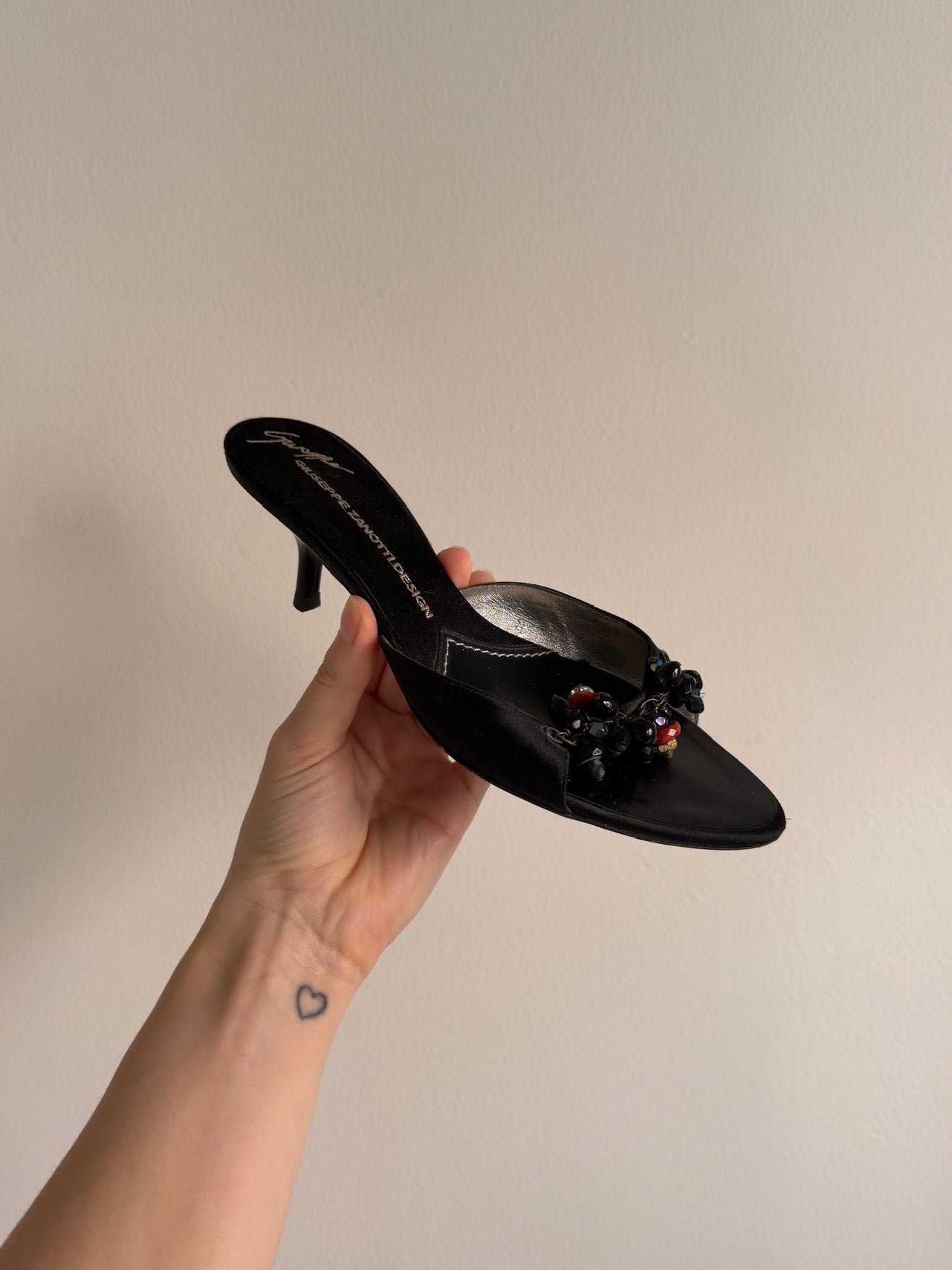 Black high-heeled shoe with decorative elements held by a hand against a beige background