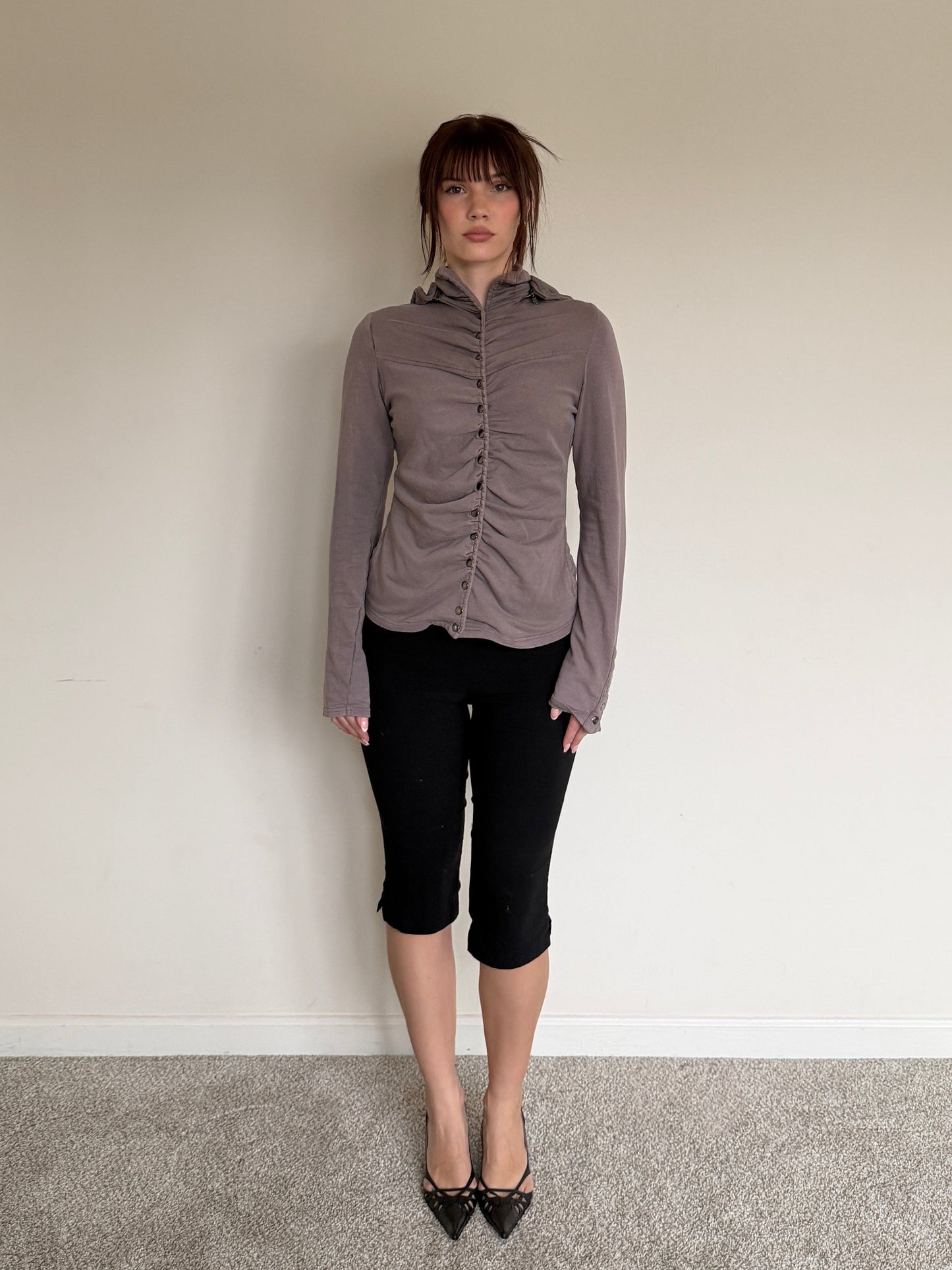 Person wearing a gray blouse and black pants standing against a plain wall.