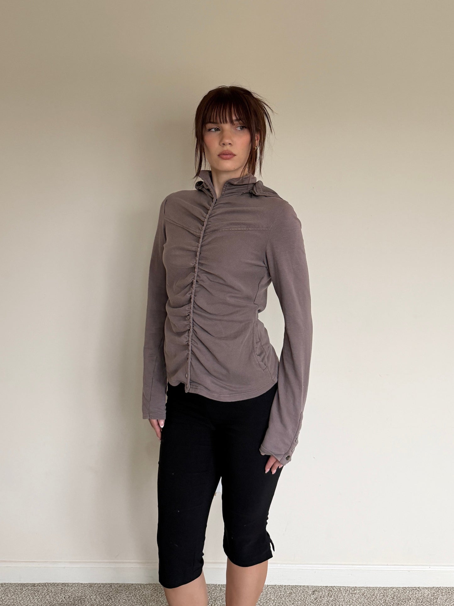 Person wearing a gray long-sleeve top and black pants against a plain wall.