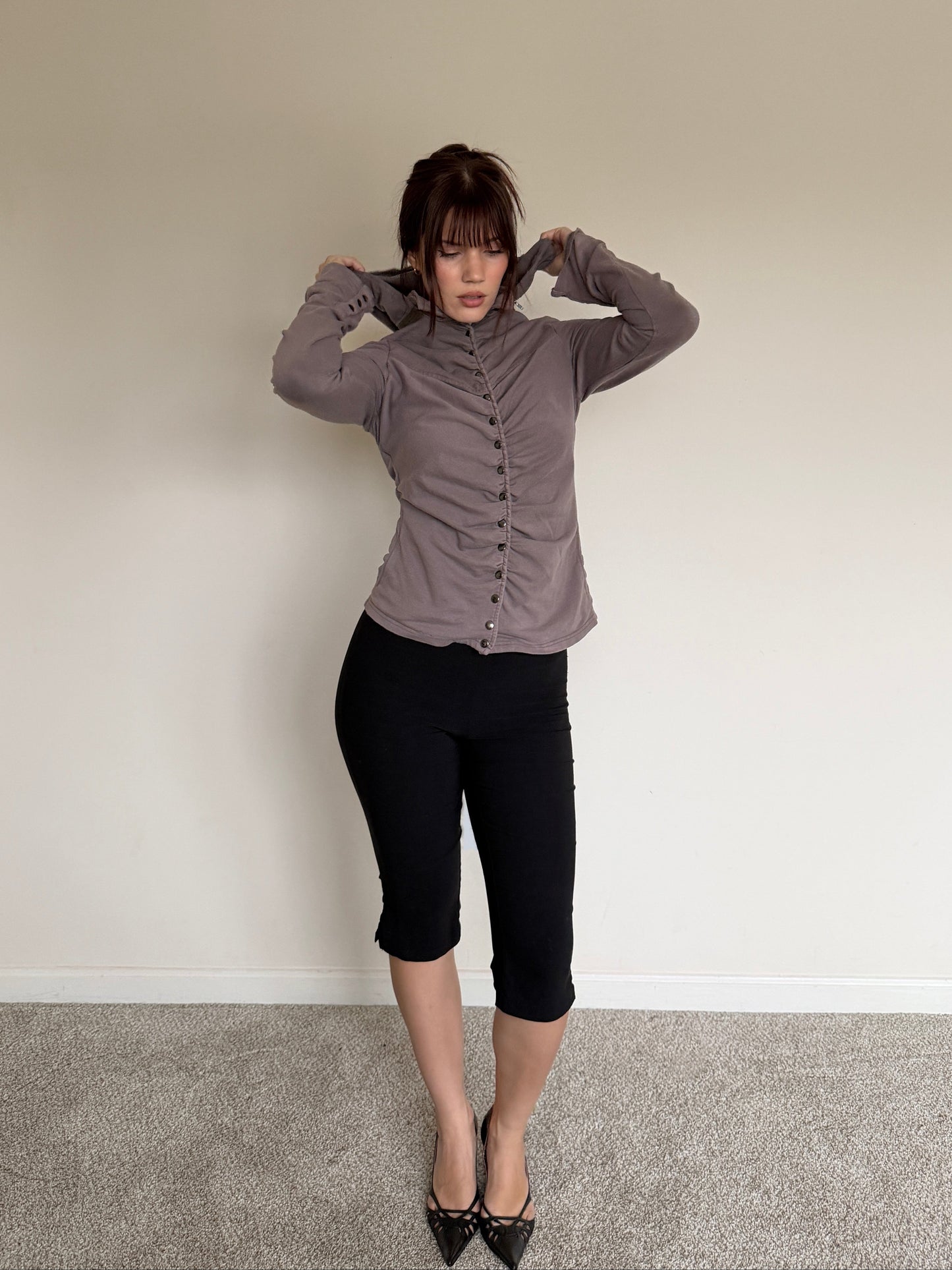 Woman wearing a gray button-up shirt and black pants standing against a plain wall.