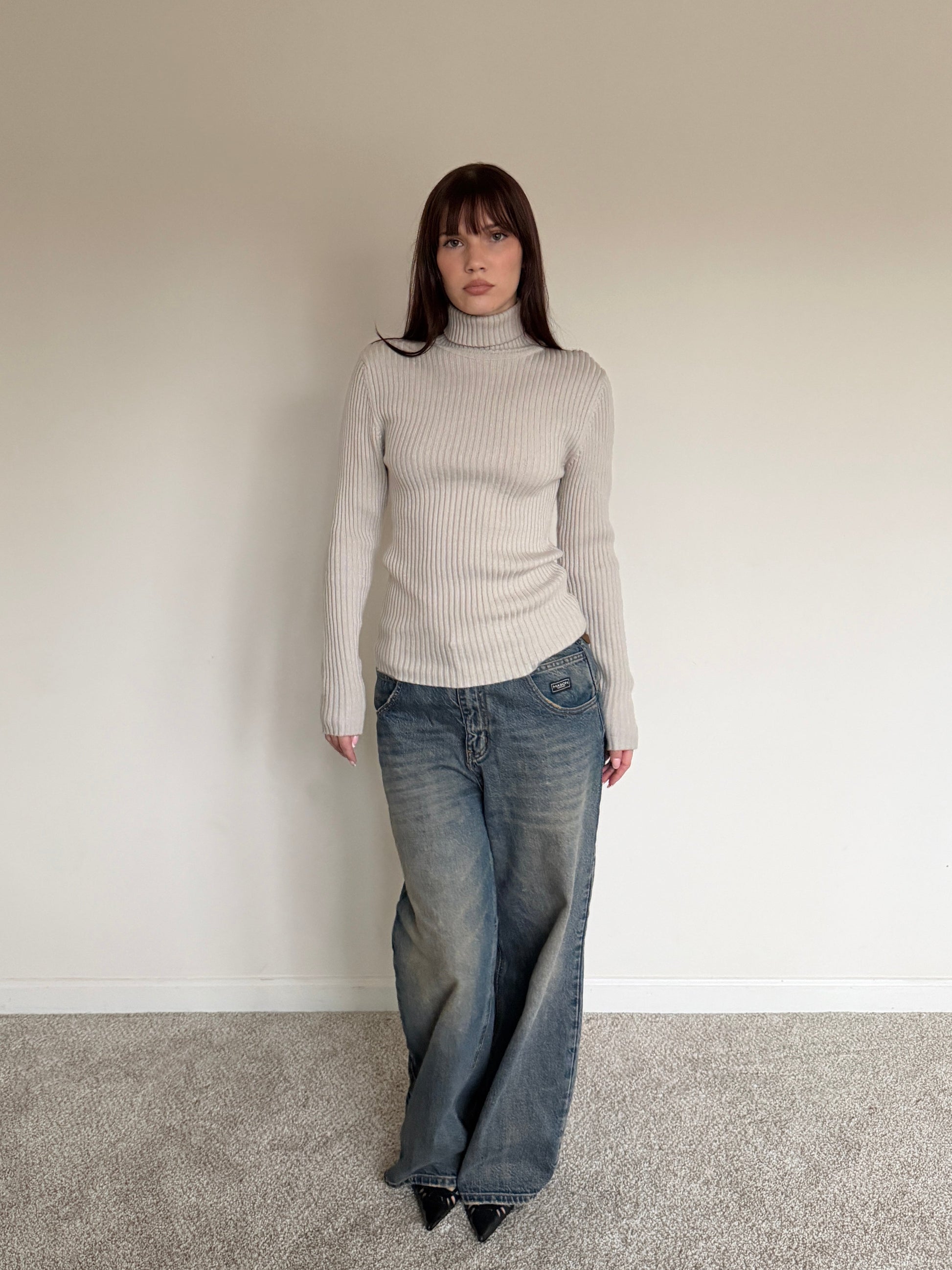 Person wearing a light gray turtleneck sweater and blue jeans against a plain wall.