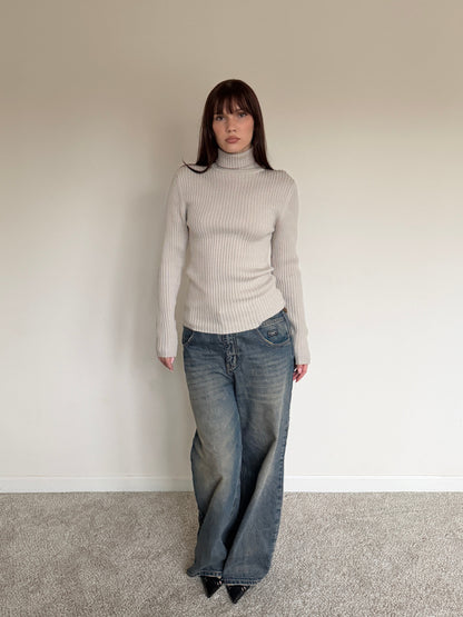 Person wearing a light gray turtleneck sweater and blue jeans against a plain wall.