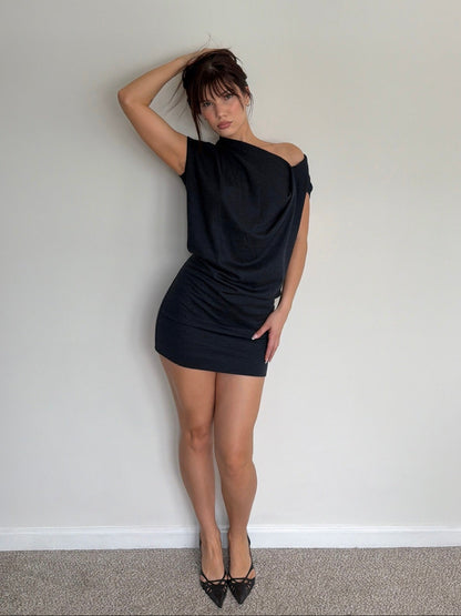 Woman wearing a black one-shoulder dress against a plain wall.