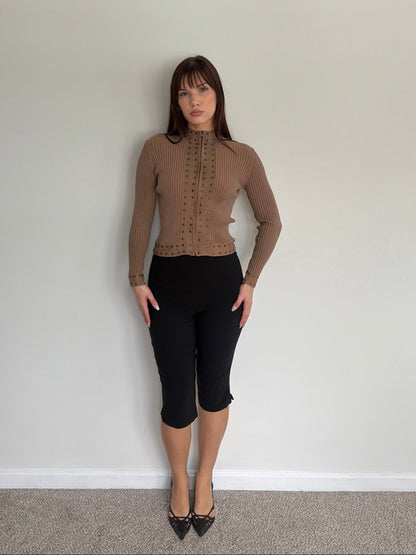 Woman wearing a brown ribbed sweater and black capri pants against a plain white wall.