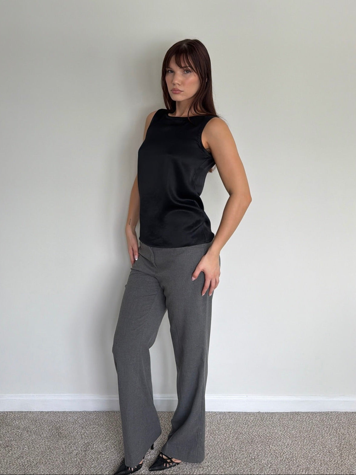 Woman wearing a black sleeveless top and gray pants against a plain wall.