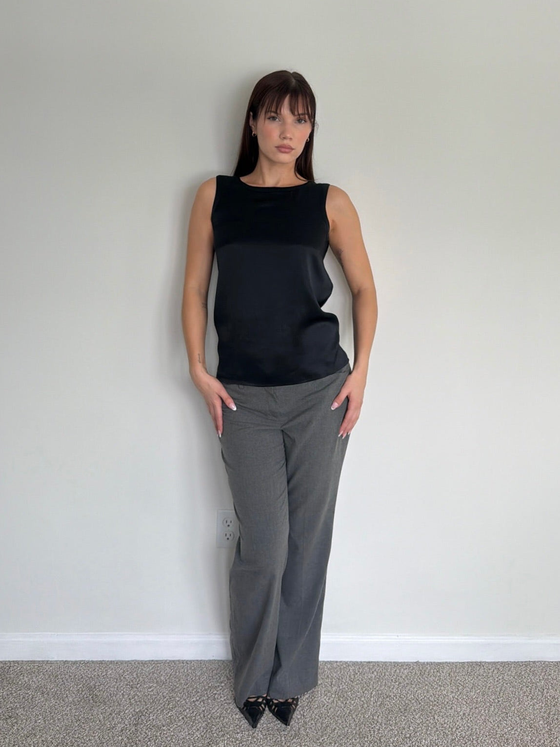 Woman wearing a black sleeveless top and gray pants against a plain white wall.