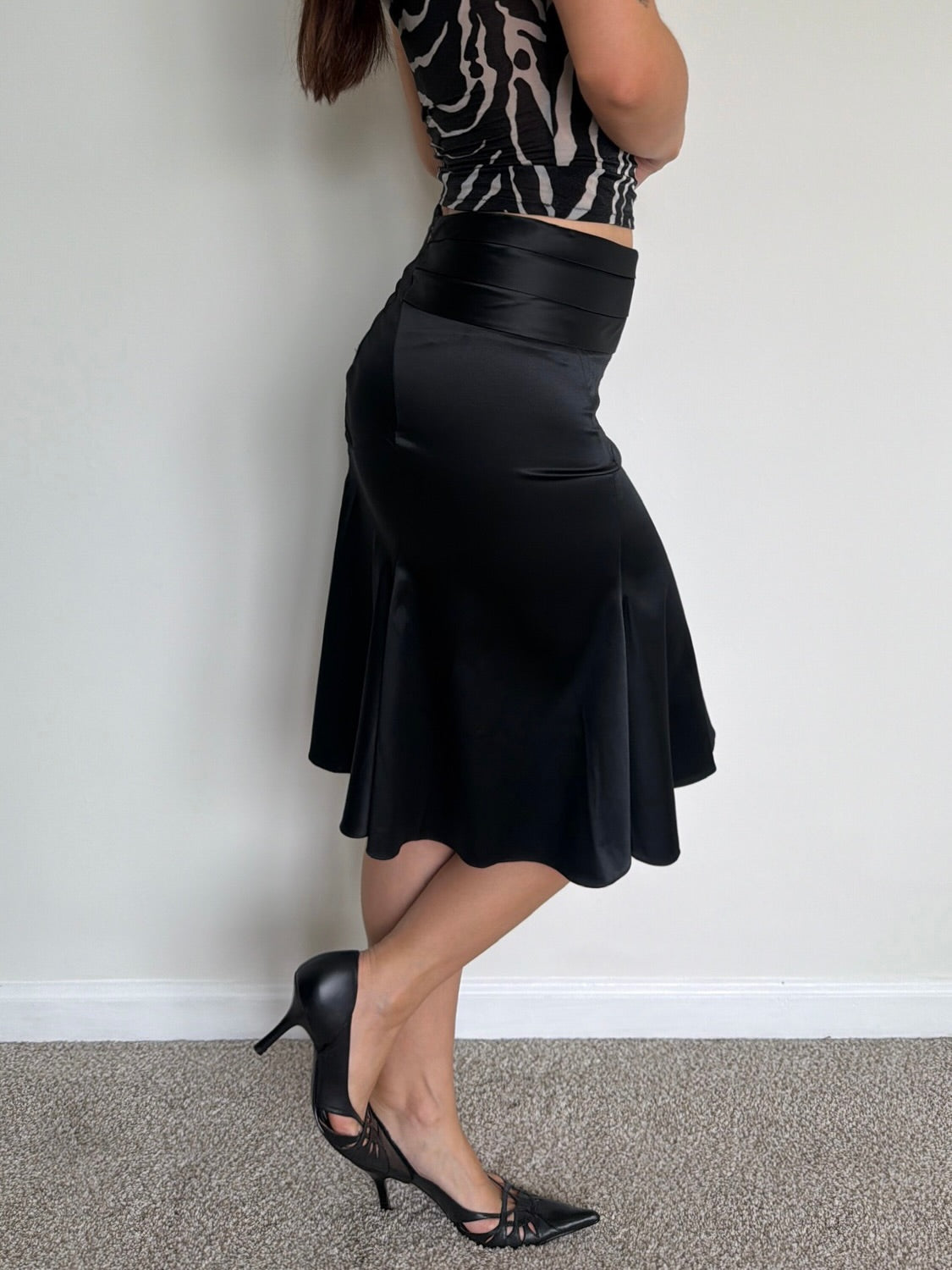 Person wearing a black skirt and high-heeled shoes against a plain wall.