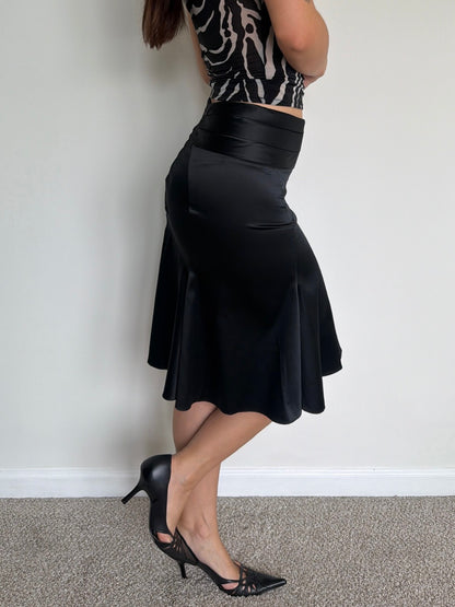 Person wearing a black skirt and high-heeled shoes against a plain wall.