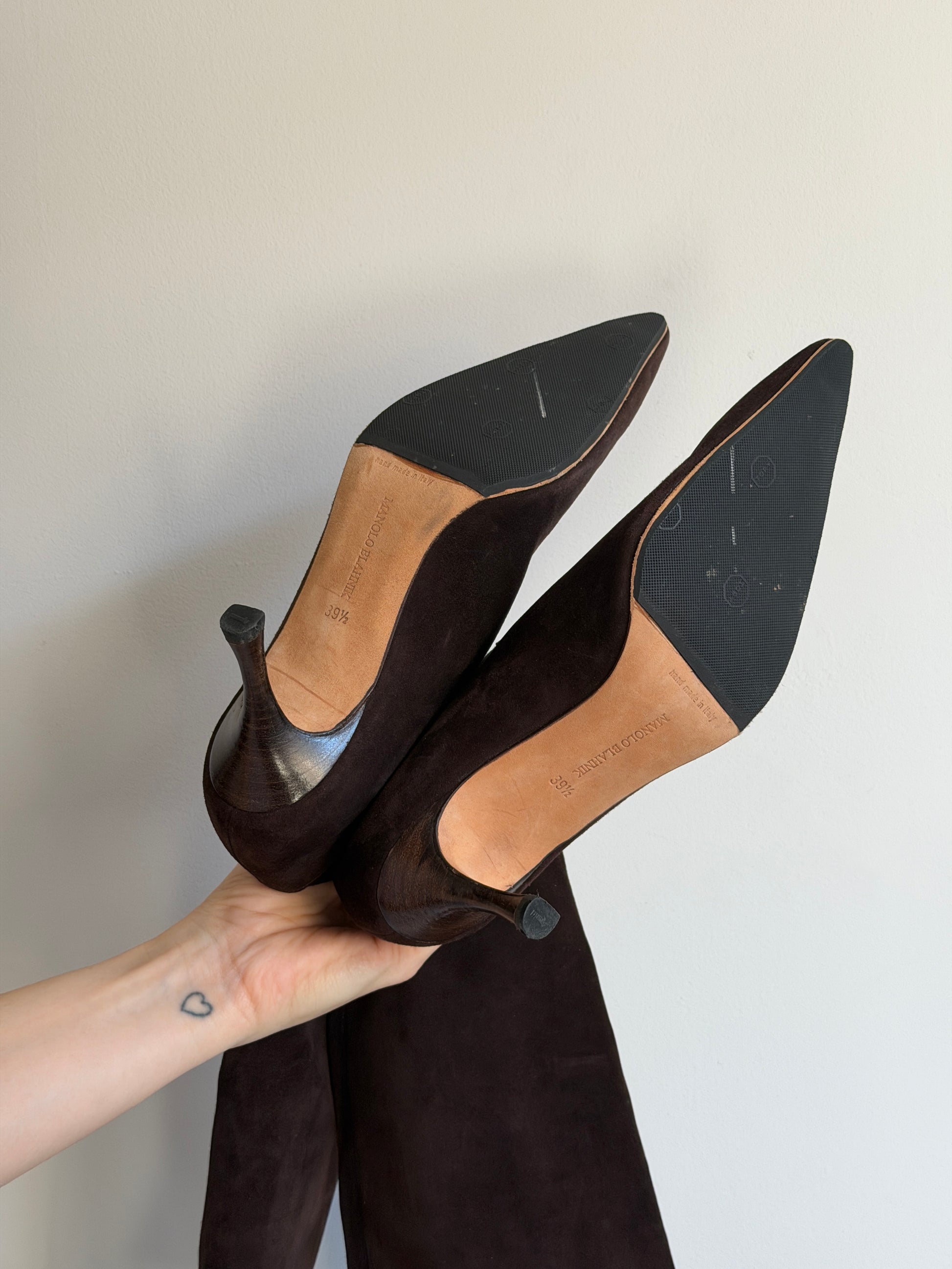 Pair of brown suede high-heeled shoes held by a hand against a plain background