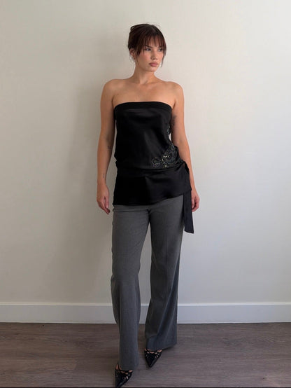 Woman wearing a black strapless top and gray pants against a plain wall.