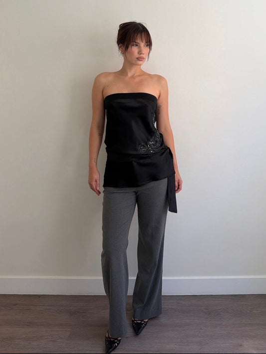 Woman wearing a black strapless top and gray pants against a plain wall.