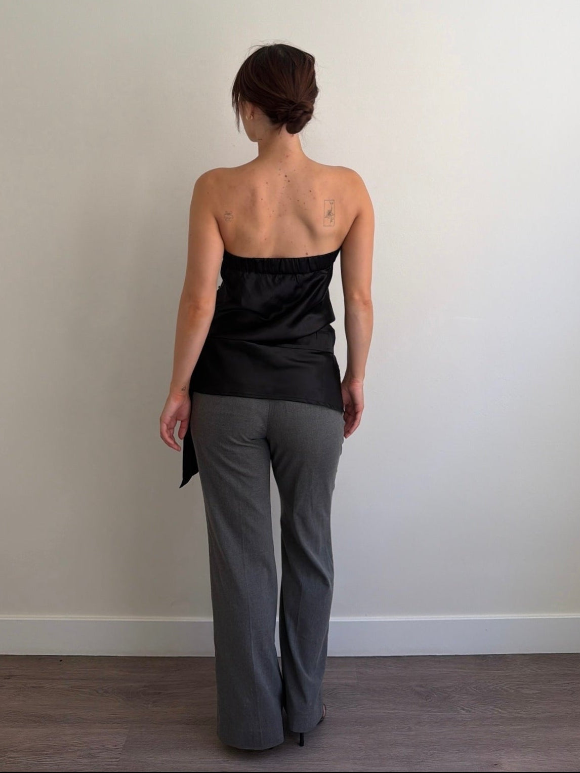 Person wearing a black strapless top and gray pants against a plain wall.