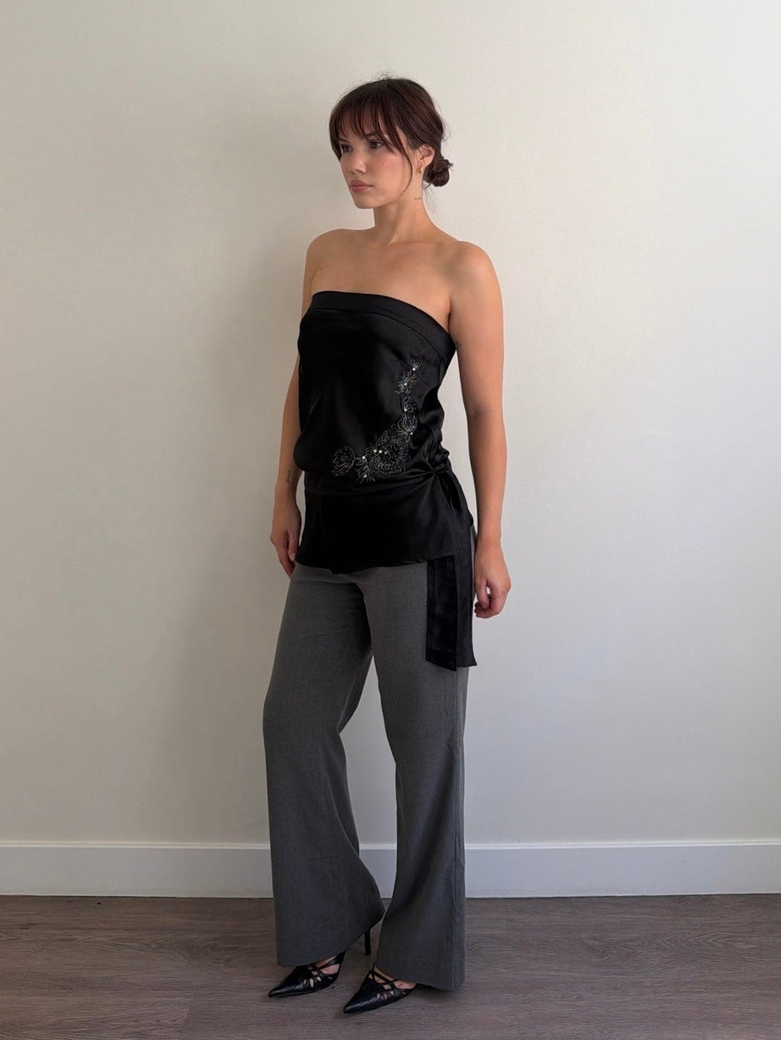 Woman wearing a black strapless top and gray pants against a plain wall.