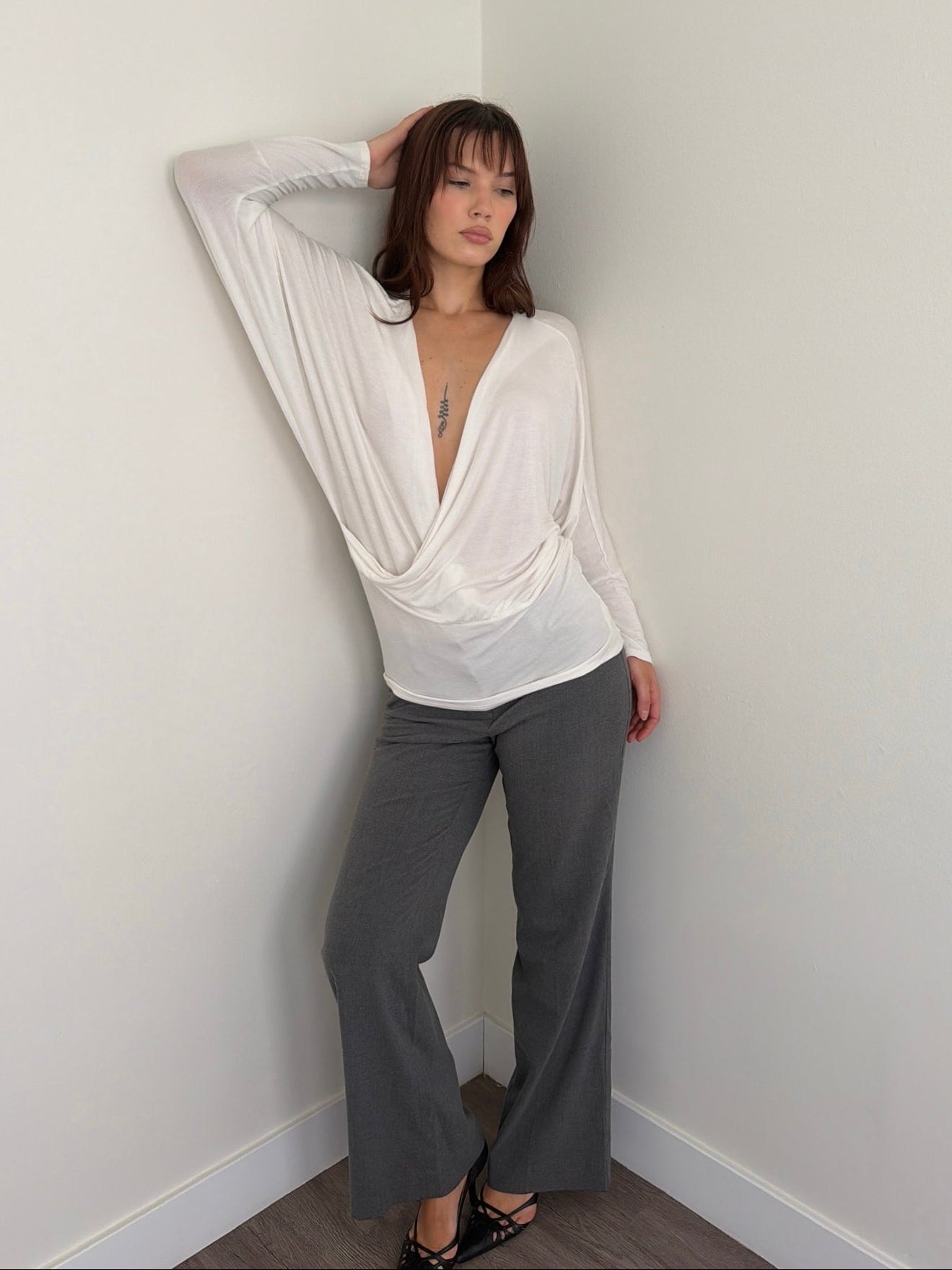 Woman wearing a white blouse and gray pants against a plain wall.