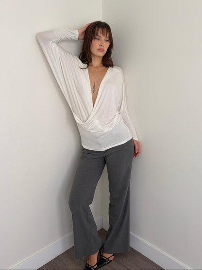 Woman wearing a white blouse and gray pants against a plain wall.