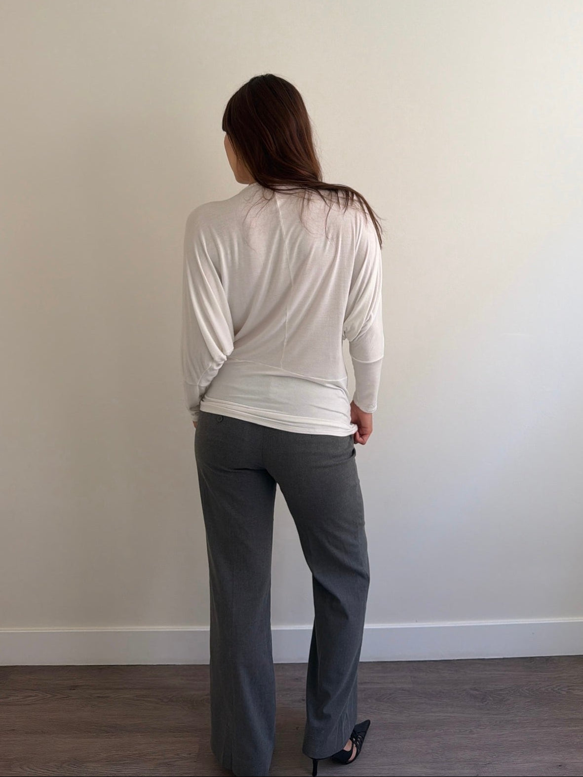 Person wearing a white top and gray pants standing against a plain wall.