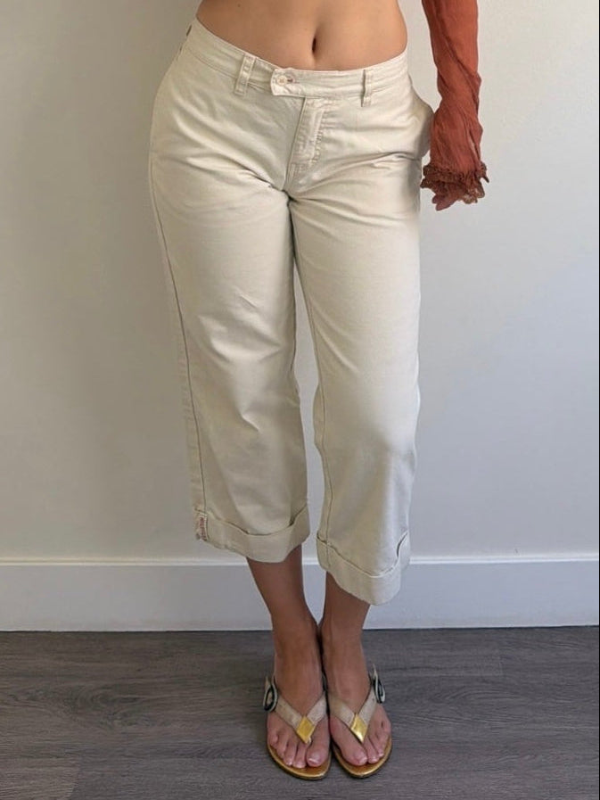 Woman wearing a brown cropped top and white pants against a plain wall.