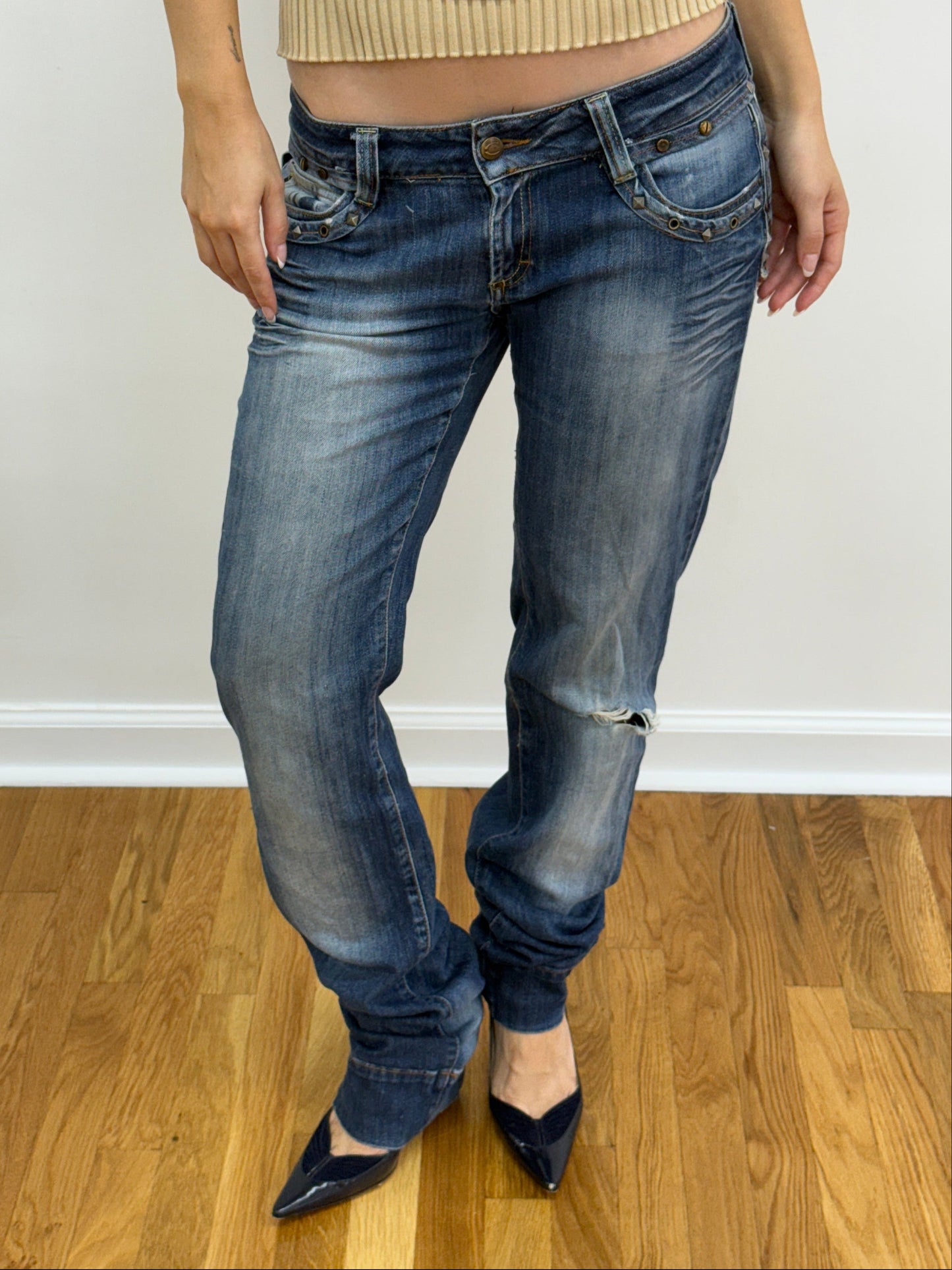 Person wearing blue jeans and black shoes on a wooden floor with a white wall background