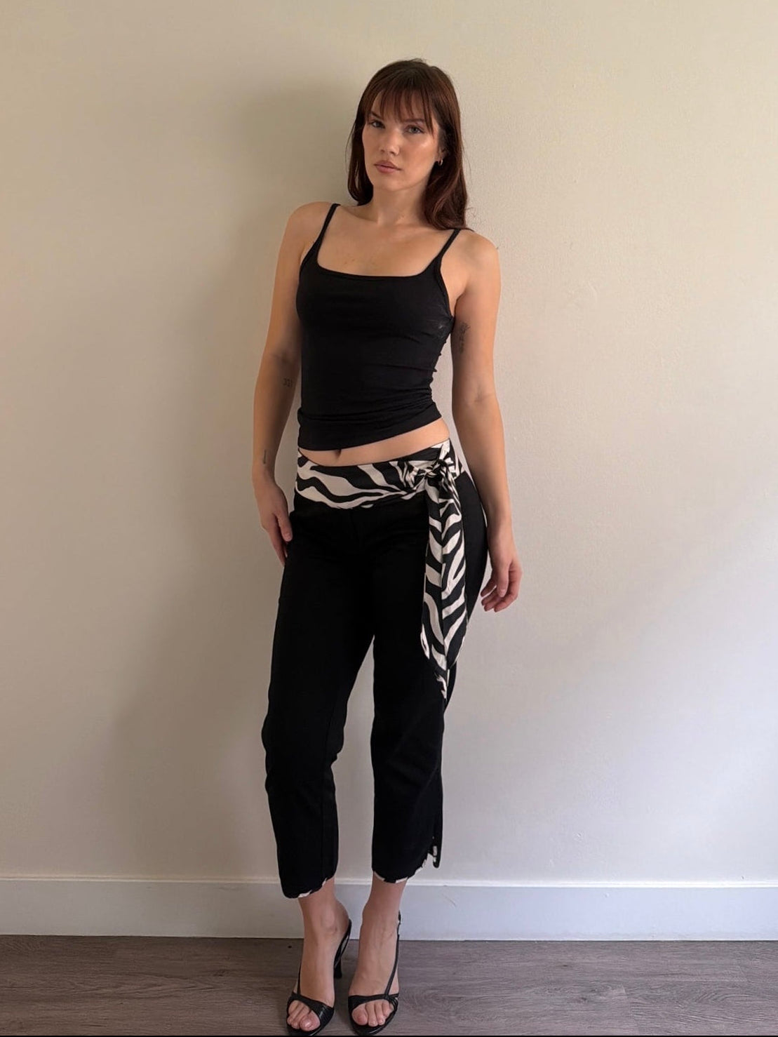 Woman wearing a black tank top and black pants with a zebra-patterned wrap against a plain wall.