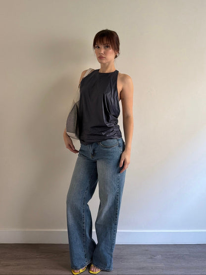 Woman wearing a dark sleeveless top and blue jeans standing against a plain wall.