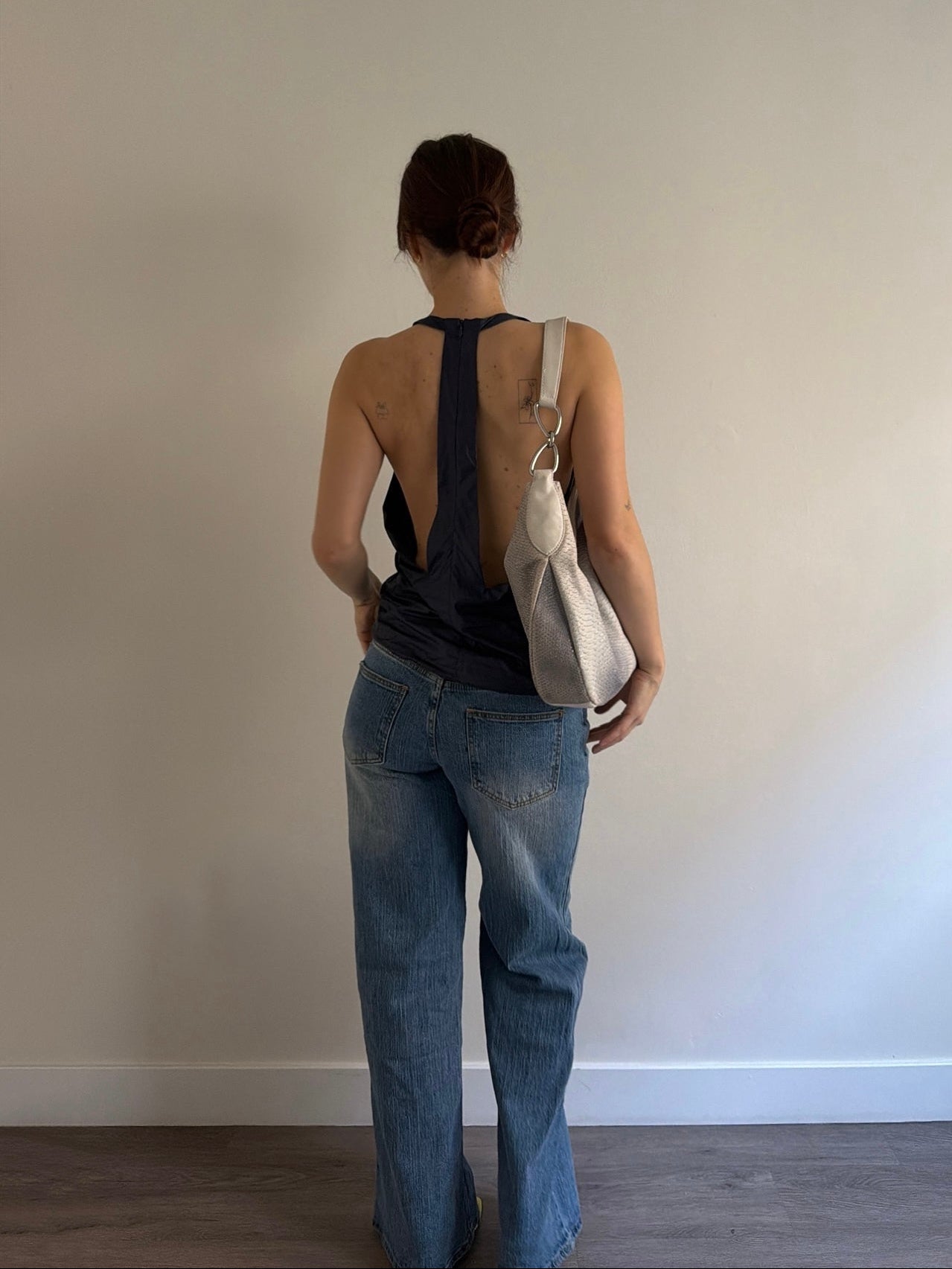 Person wearing a black sleeveless top and blue jeans, holding a beige bag against a plain wall.