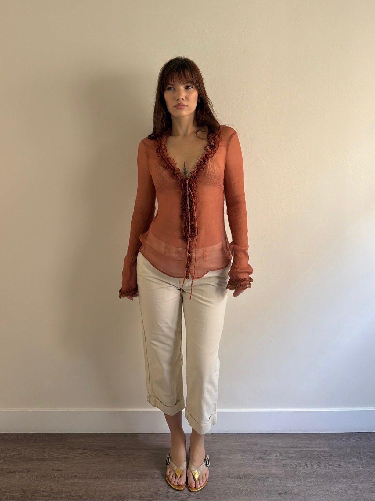 Woman wearing a brown top and beige pants standing against a plain wall.