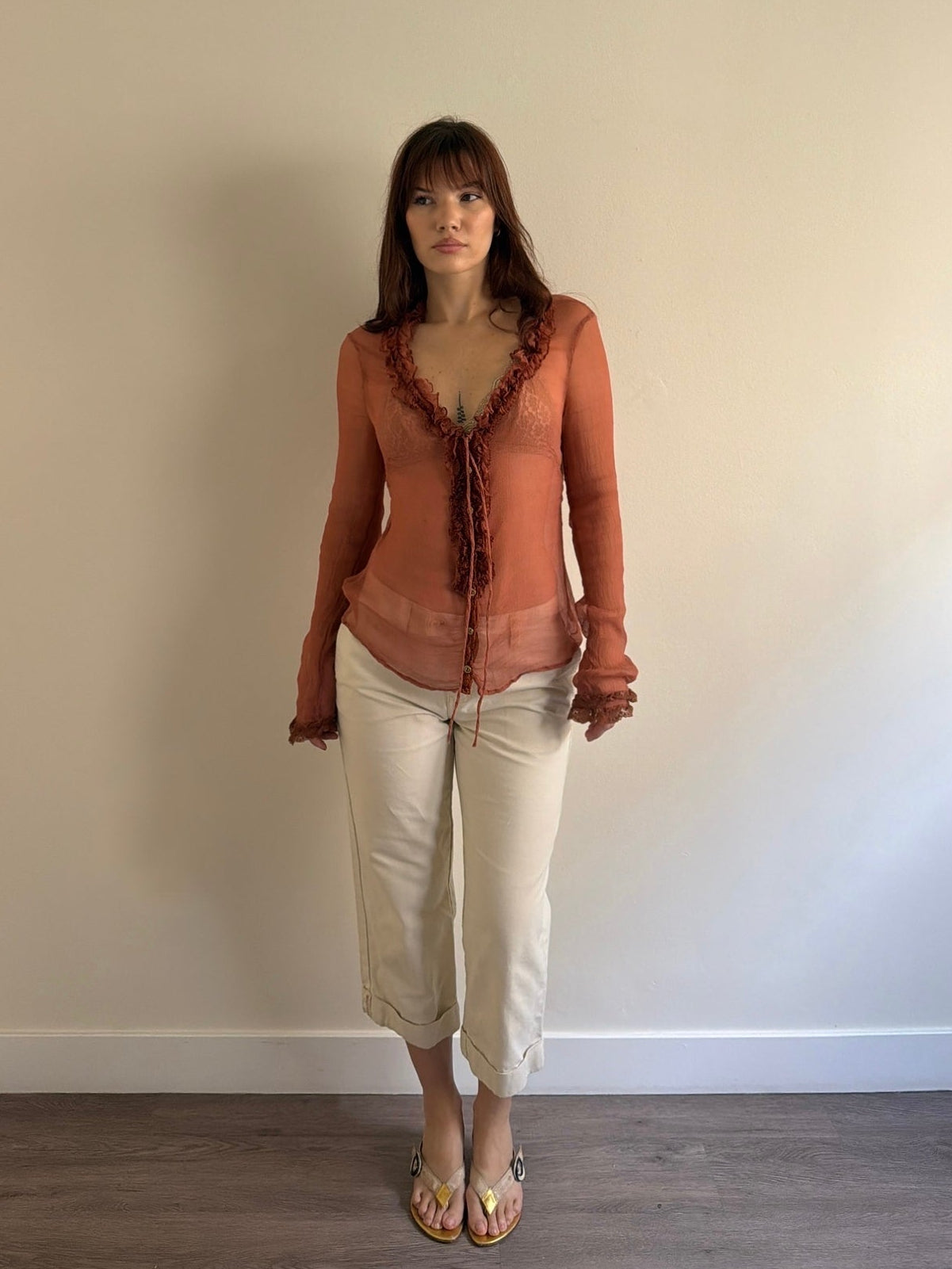 Woman wearing a brown top and beige pants standing against a plain wall.