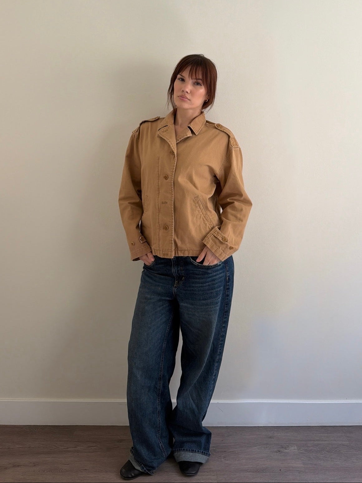 Person wearing a beige jacket and blue jeans standing against a plain wall.