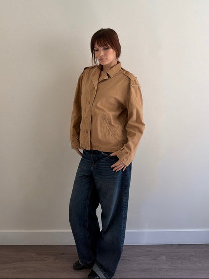 Person wearing a brown jacket and blue jeans against a plain wall.