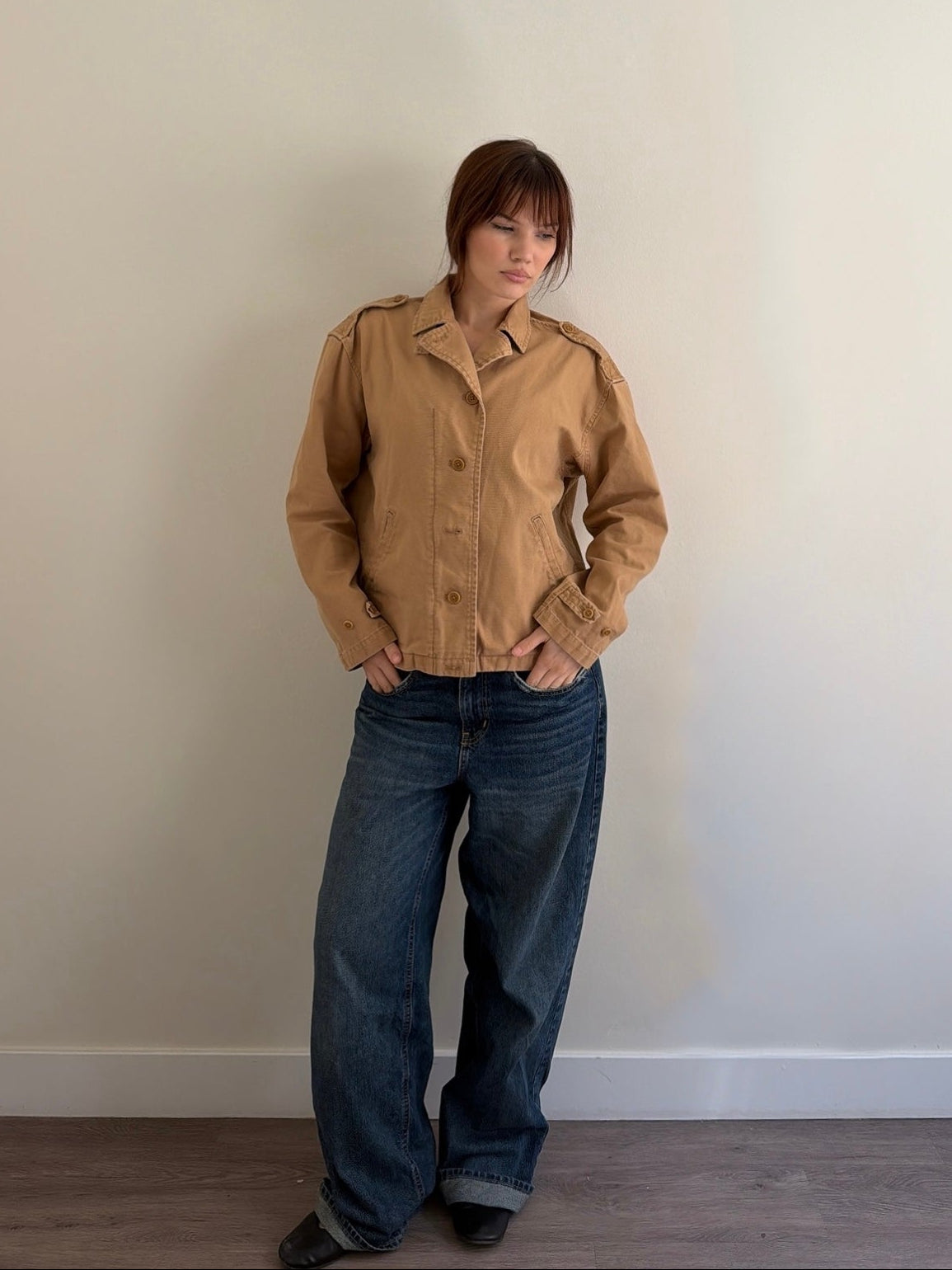 Person wearing a beige jacket and blue jeans against a plain wall.
