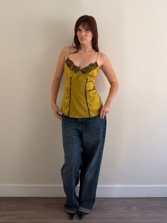 Woman wearing a mustard yellow top and blue jeans against a plain wall.
