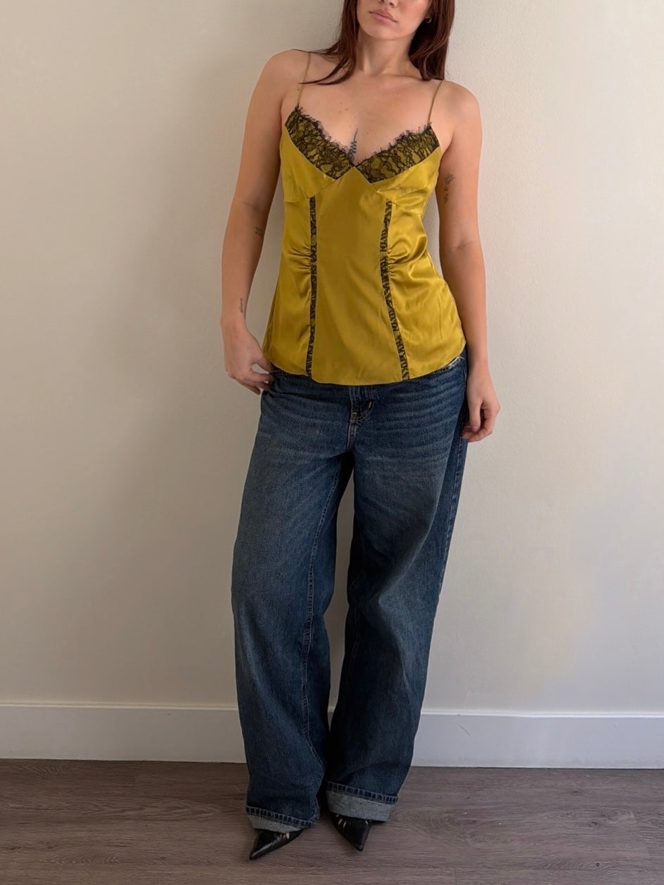Woman wearing a mustard yellow top with lace details and blue jeans against a plain wall.