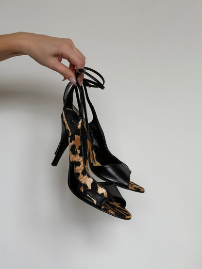 Hand holding a pair of black high-heeled shoes with animal print details against a plain background
