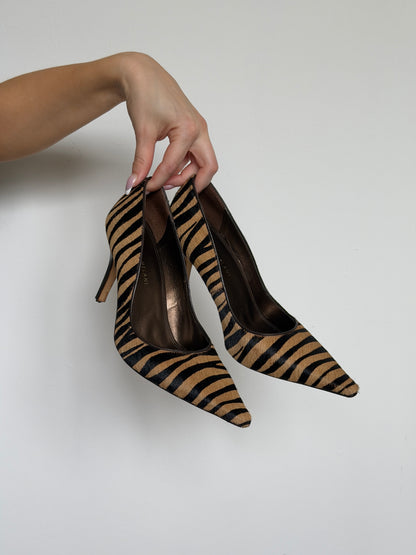 Hand holding a pair of zebra-striped high-heeled shoes against a plain background