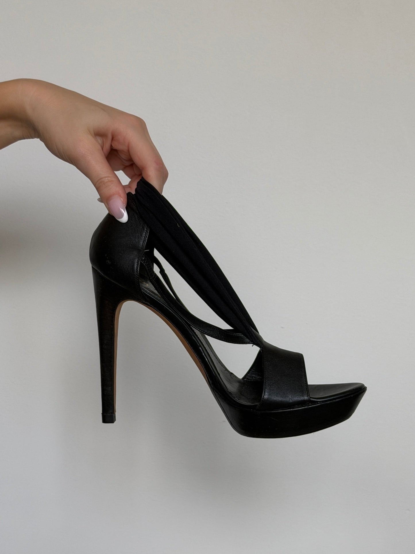 Black high-heeled shoe held by a hand against a plain background